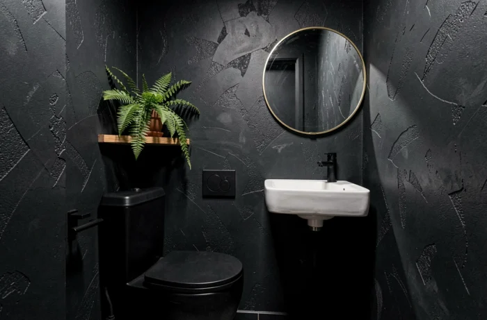 Toilettes décorées dans un contraste sombre avec des murs en anthracite mat, un miroir rond en laiton et une fougère posée sur une étagère en bois.